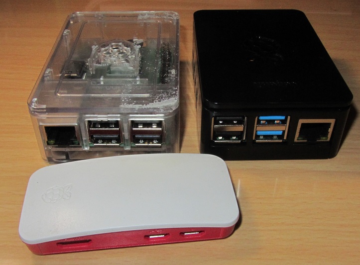 Picture showing the three Raspberry Pis being tested in their cases.  The Raspberry Pi 4B is in the black case, it is identifiable by its two blue USB 3.0 ports.